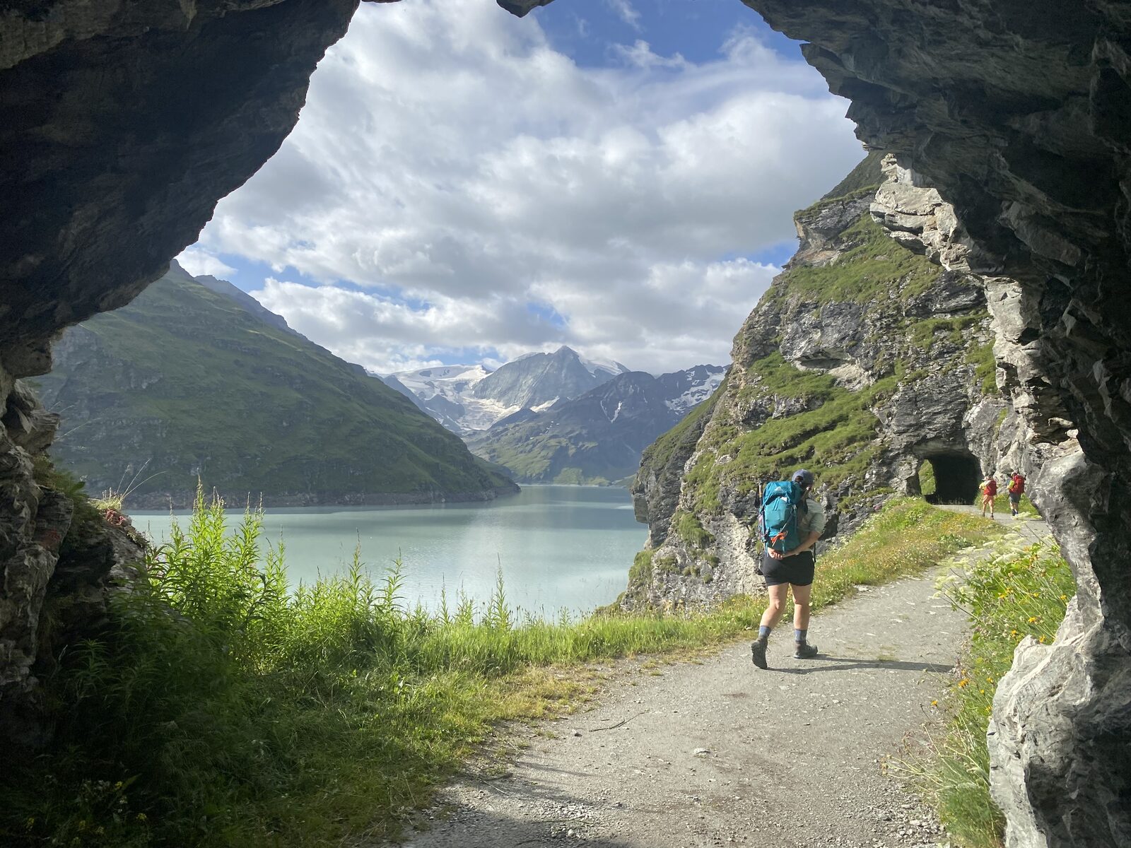 Hiker walking through a stone tunnel with a turquoise alpine lake view